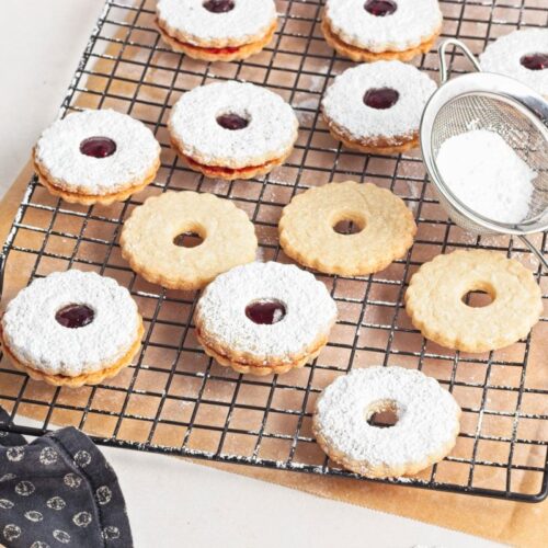 Cookies with powdered sugar and jam centers rest on a cooling rack. A sifter with powdered sugar is nearby on parchment paper, next to a dark patterned cloth.