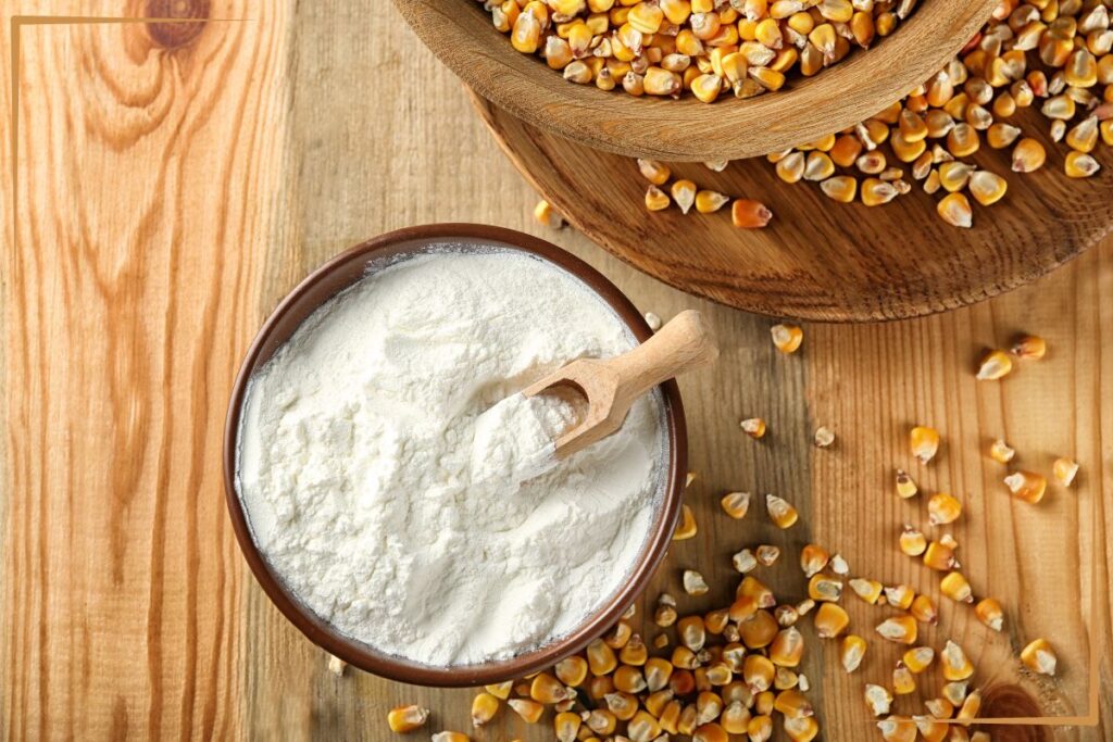 A wooden table with a bowl of white flour and a wooden scoop inside it. Next to the flour are scattered corn kernels, and a larger wooden bowl filled with more corn kernels.