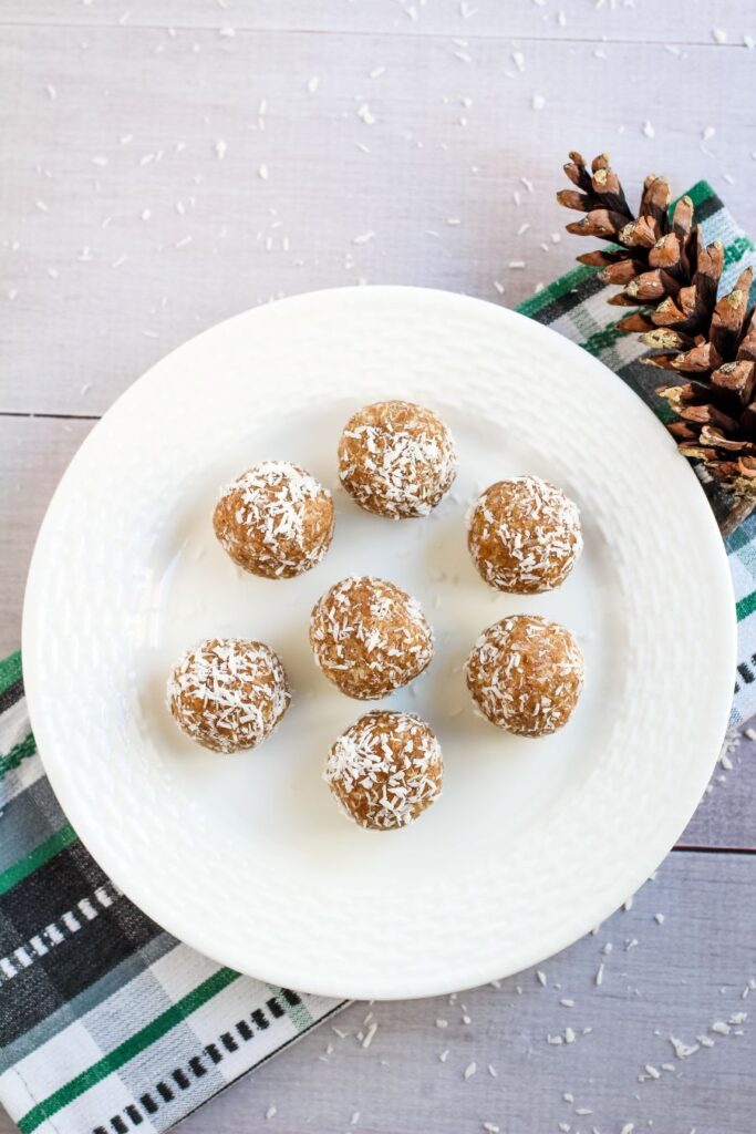A white plate with seven coconut-covered energy balls on a checkered cloth. A pinecone decorates the scene on the right, with scattered coconut flakes on the light wooden surface.