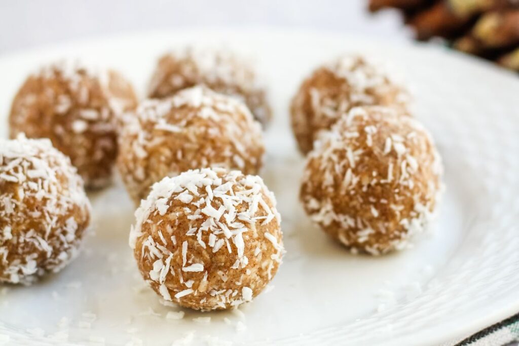 A white plate with several round energy bites covered in shredded coconut. The bites are arranged in a casual pattern, with a blurred background featuring hints of more ingredients.