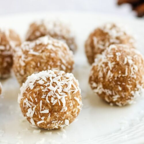 A white plate with several round energy bites covered in shredded coconut. The bites are arranged in a casual pattern, with a blurred background featuring hints of more ingredients.
