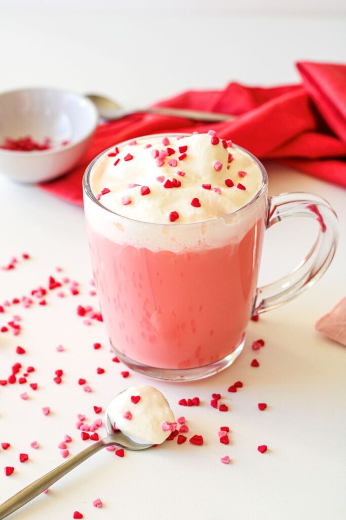 A glass mug of pink hot chocolate topped with whipped cream and red heart sprinkles. A spoon with whipped cream and sprinkles rests on a white surface, surrounded by more sprinkles. A red napkin and a small bowl are in the background.