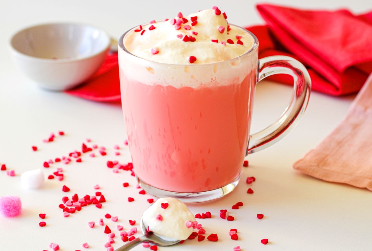 A glass mug filled with pink hot chocolate topped with whipped cream and red heart-shaped sprinkles. Beside it are scattered sprinkles, a spoon with whipped cream, a bowl, and a pink napkin in the background.