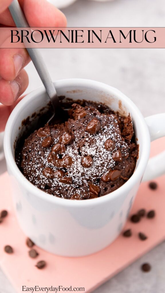 A person is holding a spoon, scooping a chocolate brownie in a mug topped with chocolate chips and powdered sugar. The mug is on a pink surface, surrounded by small chocolate chips. Text reads "Brownie in a Mug.