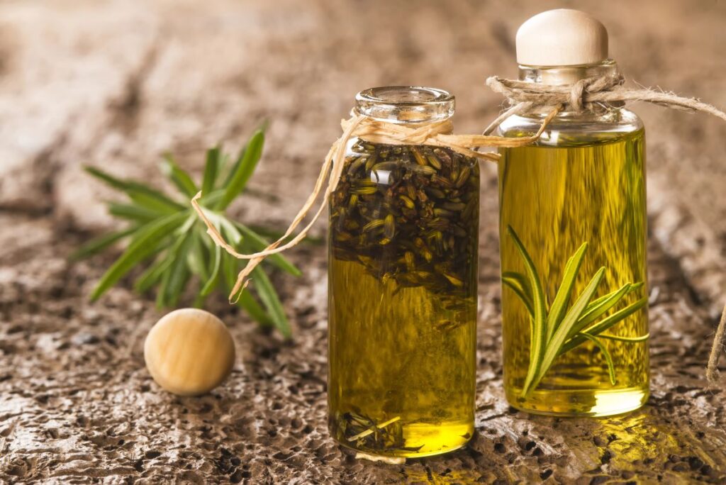 Two glass bottles filled with herbal oil sit on a rustic wooden surface. One bottle contains dark herbs, and the other has a sprig of rosemary. A round wooden stopper lies on the surface, and sprigs of greenery are in the background.