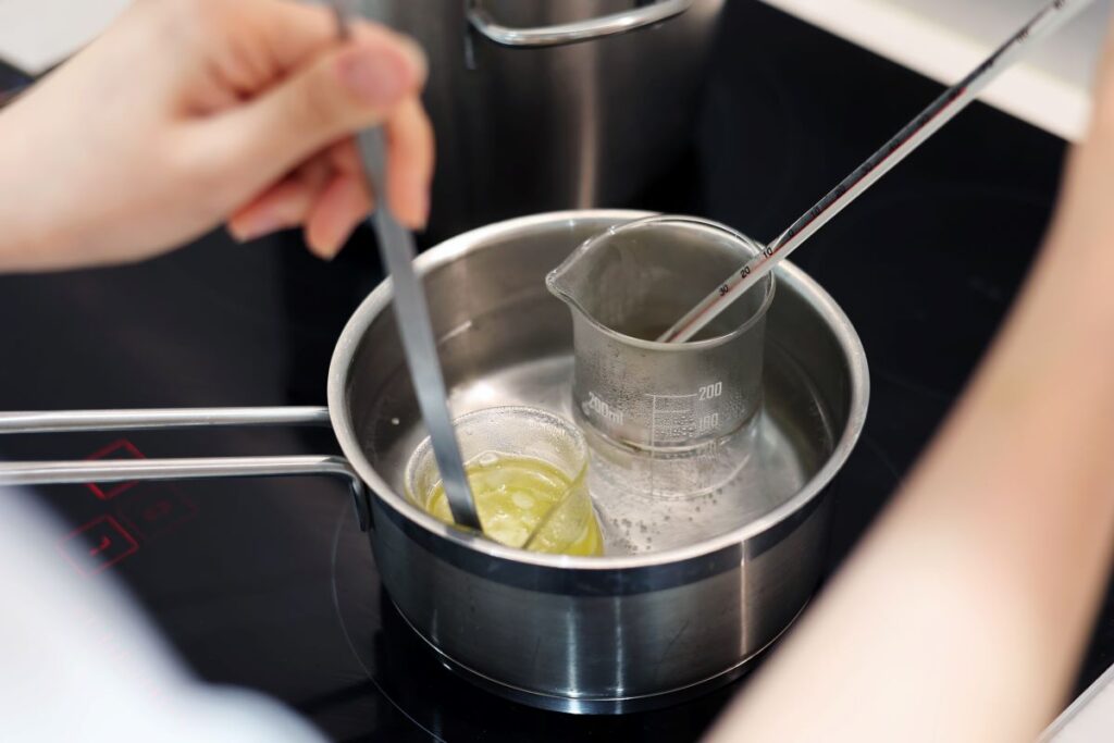 A person is holding a thermometer and a spoon over a small saucepan on a stove. Inside the saucepan, there's a beaker containing a yellow liquid, partially submerged in water, being prepared using a double boiler method.
