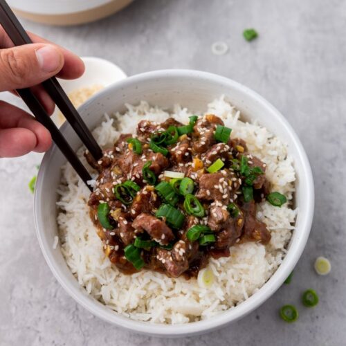A person using chopsticks to pick up a slice of beef topped with green onions and sesame seeds from a bowl of rice. The dish is on a gray surface with some sliced green onions scattered around.