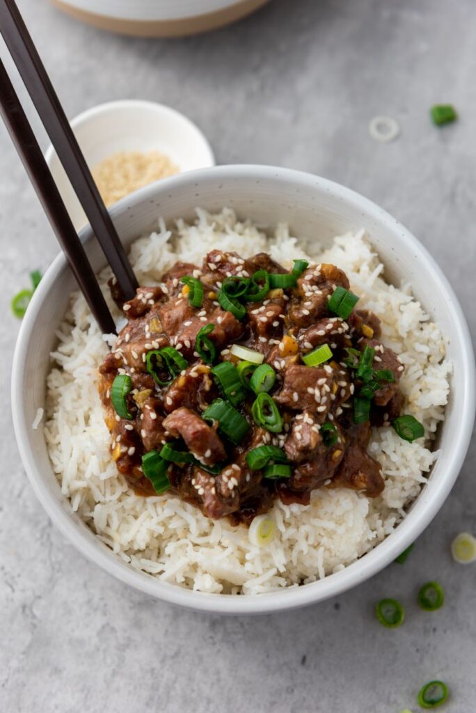 A bowl of white rice topped with beef in a savory sauce, garnished with chopped green onions and sesame seeds. Two chopsticks rest on the side of the bowl. A small bowl of sesame seeds is in the background.