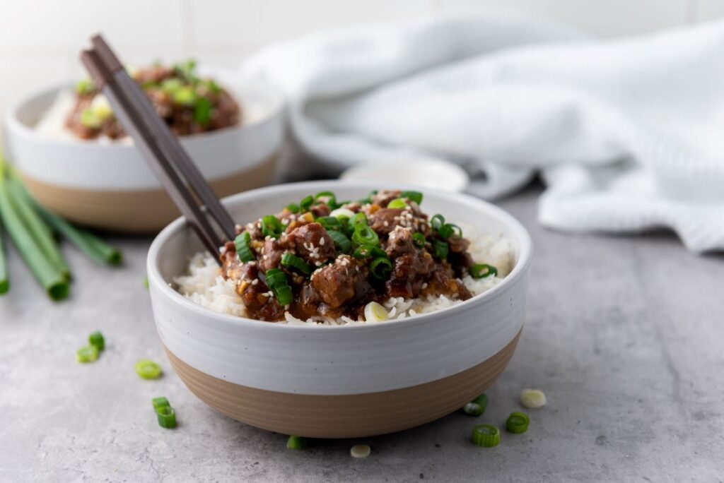 A bowl of rice topped with a savory meat dish garnished with chopped green onions and sesame seeds. A pair of chopsticks rests on the bowl. Another similar bowl is blurred in the background on a gray surface. Scallions are scattered beside the bowl.