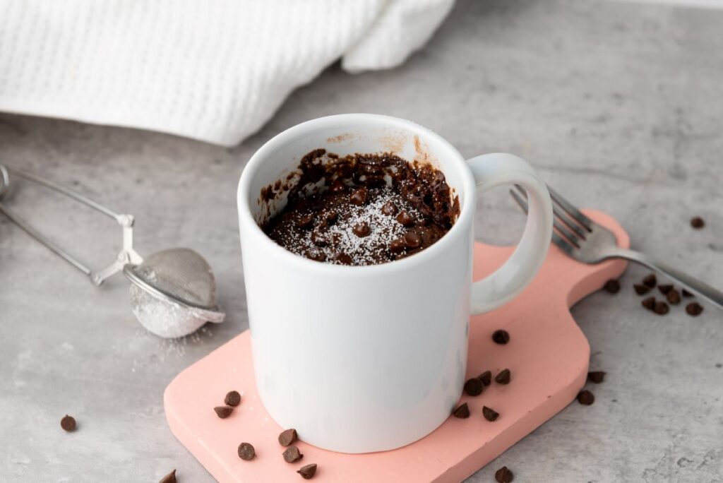 A white mug filled with chocolate cake sits on a pink cutting board. Chocolate chips are scattered around. A dusting of powdered sugar and a small mesh strainer are visible. A fork and white cloth are in the background on a gray surface.