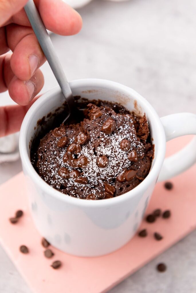 A hand holds a spoon inside a mug filled with chocolate cake topped with powdered sugar and chocolate chips. The mug sits on a pink rectangular surface with scattered coffee beans.