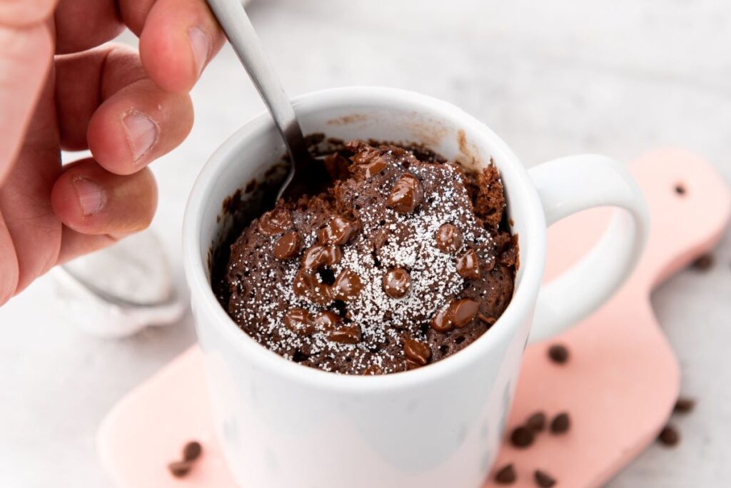A hand holding a spoon is about to scoop from a chocolate mug cake topped with powdered sugar and chocolate chips. The mug is white and placed on a light pink cutting board, with scattered chocolate chips around.