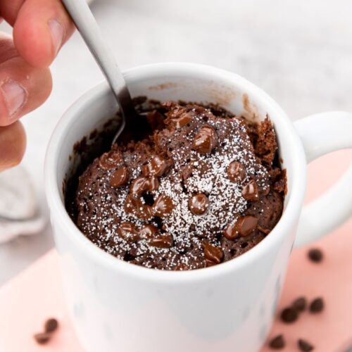 A hand holding a spoon is about to scoop from a chocolate mug cake topped with powdered sugar and chocolate chips. The mug is white and placed on a light pink cutting board, with scattered chocolate chips around.
