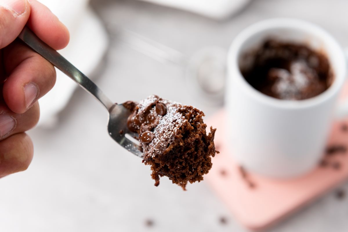 Close-up of a hand holding a spoonful of chocolate cake with powdered sugar. In the background, there's a white mug with more chocolate cake, placed on a light surface.