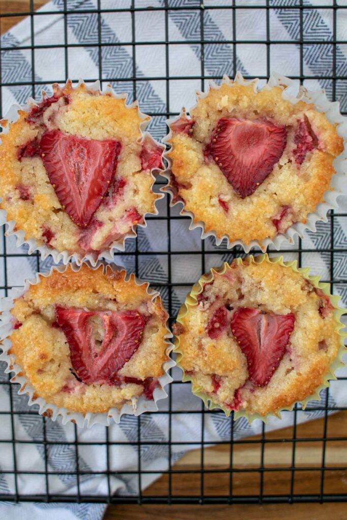 Four strawberry muffins topped with sliced strawberries sit on a cooling rack placed over a checkered cloth.