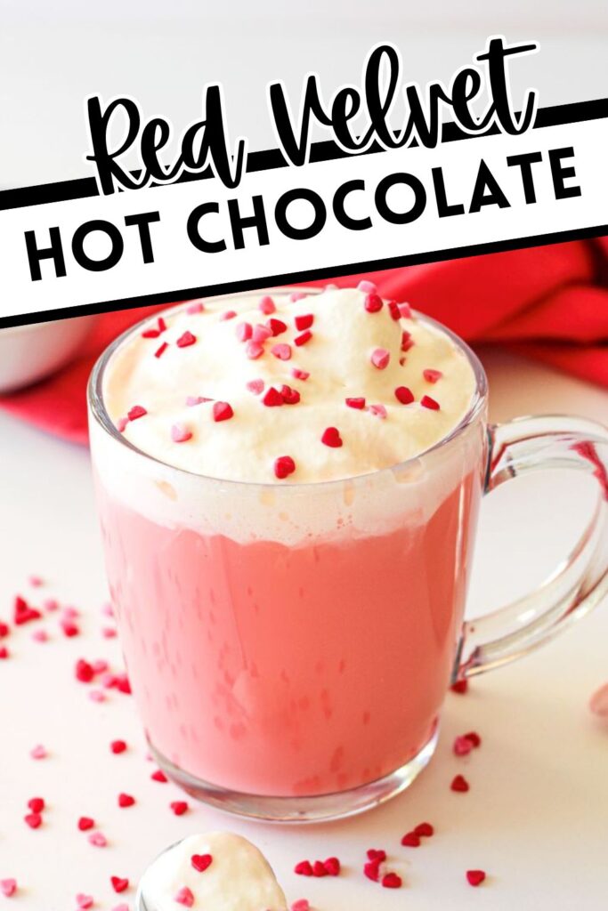 A clear glass mug filled with red velvet hot chocolate topped with whipped cream and small red heart sprinkles. The text above the mug reads "Red Velvet Hot Chocolate." Scattered heart sprinkles are visible around the mug.