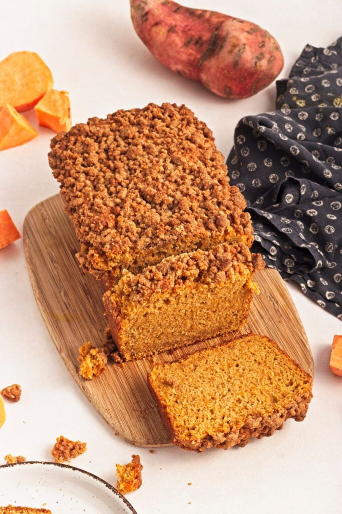 A loaf of sweet potato bread with crumble topping, partially sliced, on a wooden board. A whole sweet potato and fabric with a pattern are in the background. Slices of sweet potato are scattered around.