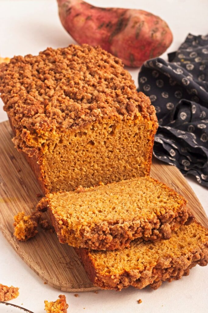 A loaf of sweet potato bread on a wooden board, with two slices cut. The bread has a crumbly, golden brown crust. A sweet potato and a dark cloth with white patterns are in the background.