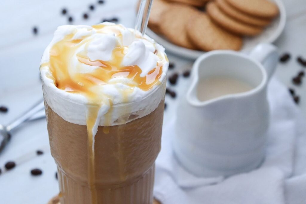 A tall glass of iced coffee topped with whipped cream and caramel sauce, next to a small white pitcher of cream. In the background, a plate of cookies is slightly out of focus, with scattered coffee beans on the table.