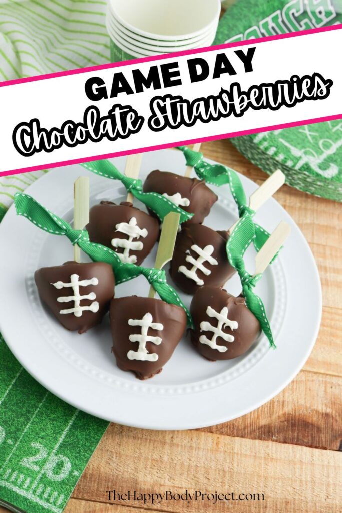 A white plate holds chocolate-covered strawberries decorated to look like footballs with white icing laces. Each strawberry has a small clothespin and a green ribbon attached. A stack of paper cups is in the background on a wooden table.