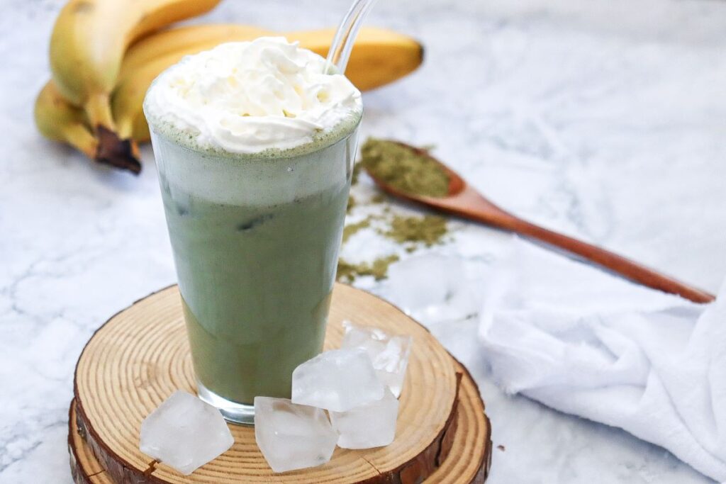 A glass of green tea latte topped with whipped cream and a straw sits on a wooden coaster. Ice cubes are scattered around. In the background, bananas and a wooden spoon with matcha powder are visible on a marble surface.