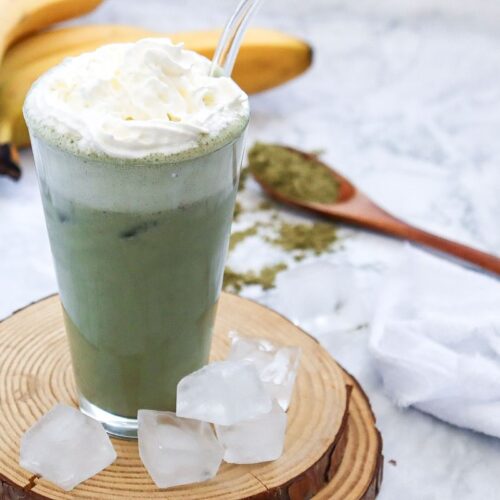 A glass of green tea latte topped with whipped cream and a straw sits on a wooden coaster. Ice cubes are scattered around. In the background, bananas and a wooden spoon with matcha powder are visible on a marble surface.