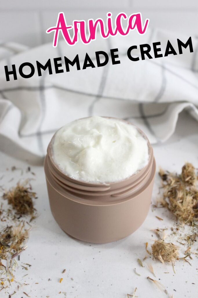 A small jar filled with white homemade arnica cream is placed on a white surface. Dried arnica flowers are scattered around the jar. A white and gray checkered cloth is in the background. The text "Arnica Homemade Cream" is displayed above.