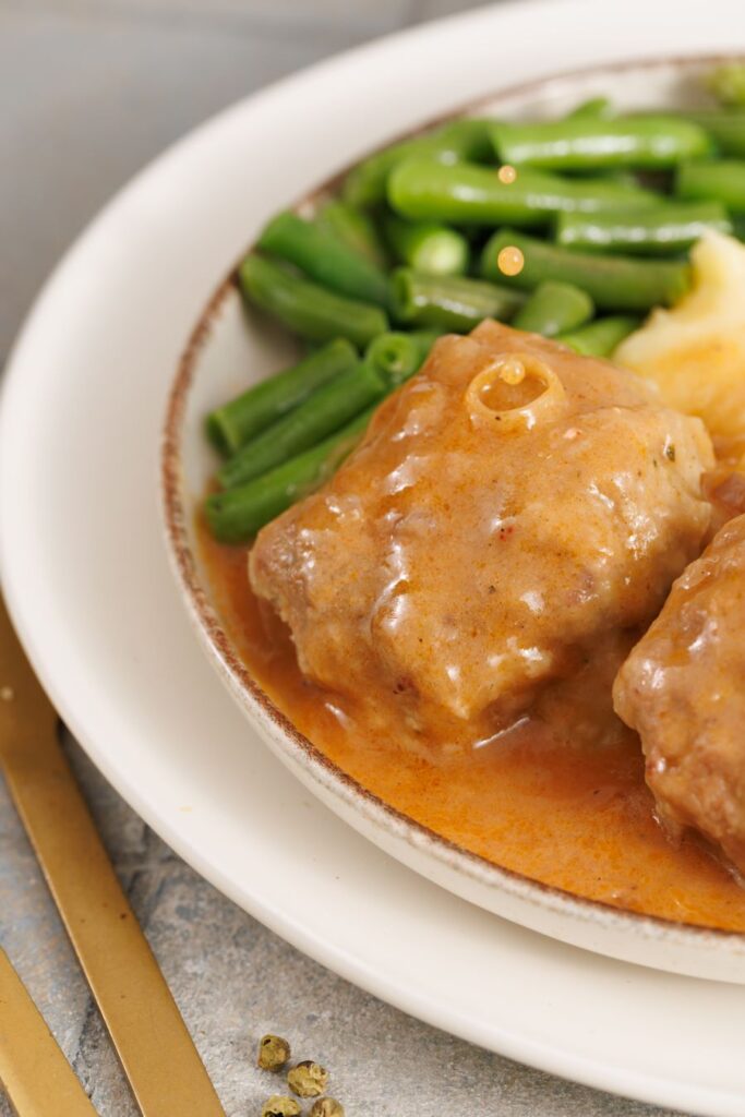 A plate of meat with gravy, accompanied by mashed potatoes and green beans. The meal is served on a white ceramic plate with a light brown rim, placed on a gray surface. A gold fork is visible beside the plate.