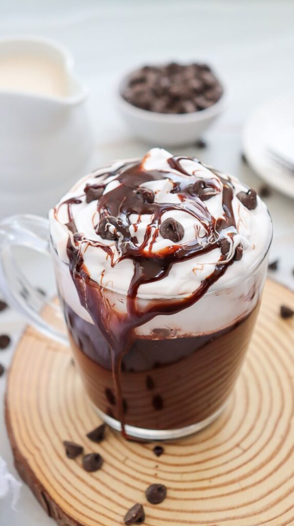 A glass mug filled with hot chocolate topped with whipped cream, drizzled with chocolate syrup, and sprinkled with chocolate chips. It rests on a wooden coaster, with more chocolate chips scattered around. A jug and a bowl are blurred in the background.