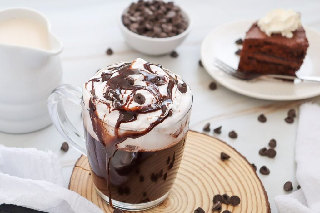 A glass mug filled with hot chocolate topped with whipped cream and chocolate drizzle sits on a wooden coaster. In the background, there's a slice of chocolate cake on a plate, a bowl of chocolate chips, and a white pitcher.