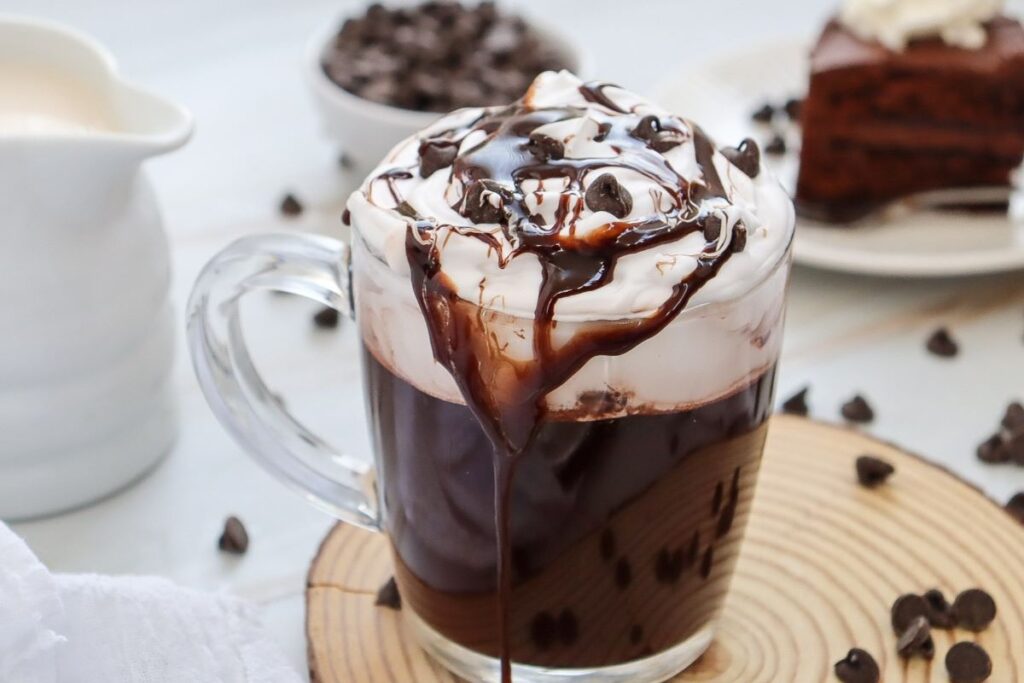 A glass mug filled with rich dark hot chocolate is topped with whipped cream, chocolate syrup, and chocolate chips. It sits on a wooden coaster with chocolate chips scattered around. A blurred chocolate cake slice is in the background.