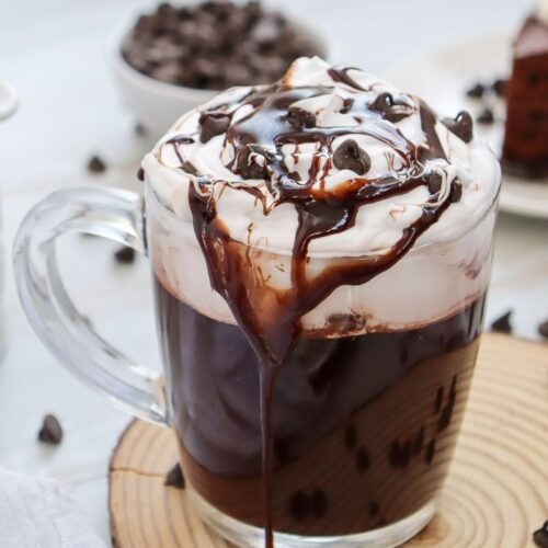 A glass mug filled with rich dark hot chocolate is topped with whipped cream, chocolate syrup, and chocolate chips. It sits on a wooden coaster with chocolate chips scattered around. A blurred chocolate cake slice is in the background.