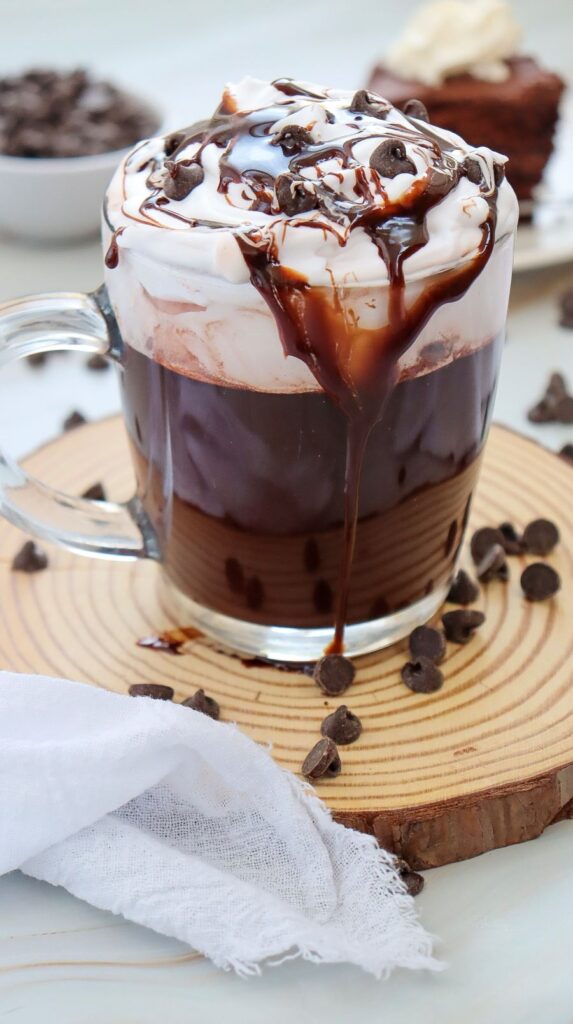A glass mug filled with hot chocolate, topped with whipped cream, chocolate drizzle, and chocolate chips. It sits on a wooden coaster, surrounded by scattered chocolate chips and a white cloth. A dessert is blurred in the background.