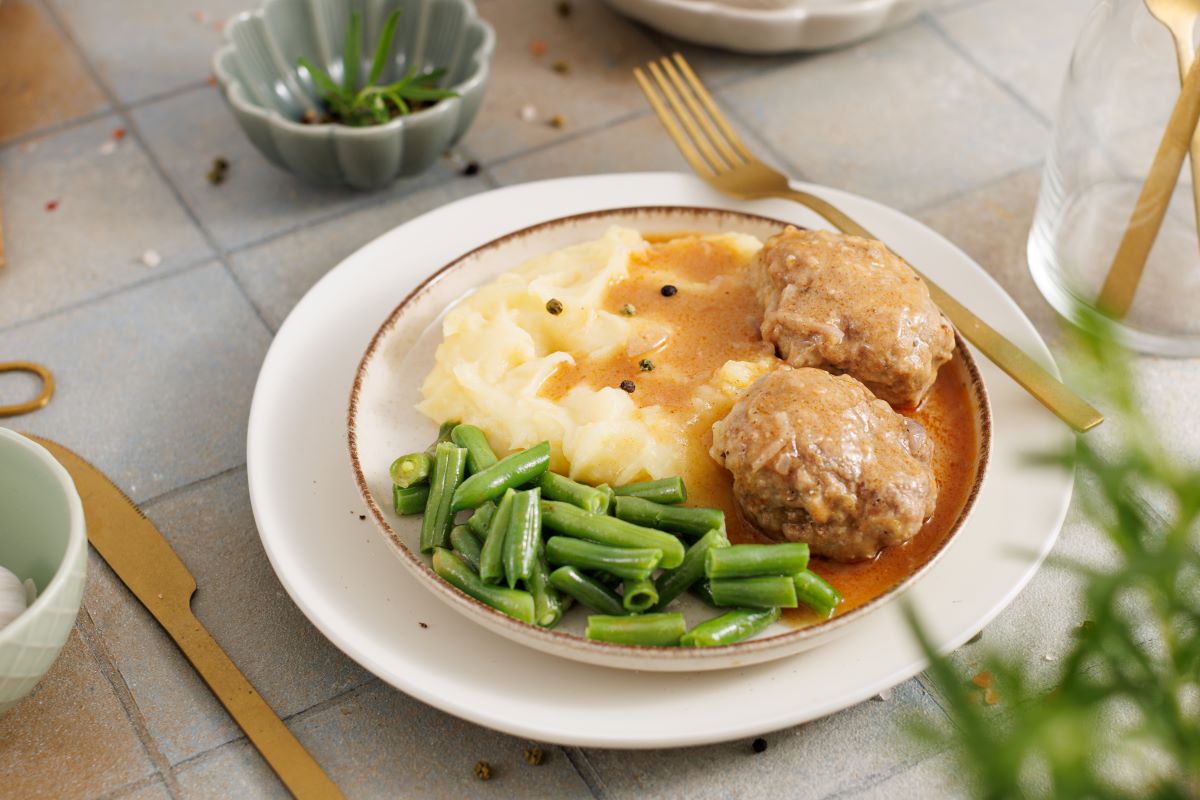 A plate of mashed potatoes, meatballs in gravy, and green beans is served on a table with a gold fork and knife. A small bowl of herbs and ingredients is nearby, set on a tiled surface with scattered peppercorns.