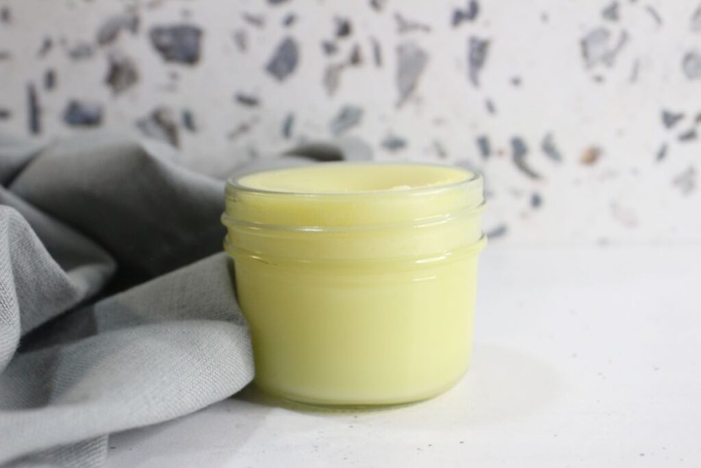 A small glass jar filled with a smooth, yellow balm sits on a white surface. A soft, gray cloth is partially visible to the left, and the background features a white wall with a terrazzo pattern of scattered gray and black flecks.