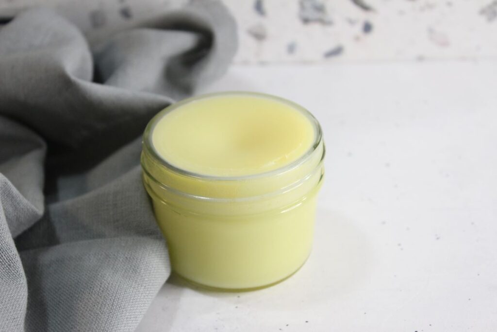 A small glass jar filled with pale yellow cream sits on a light surface. A soft gray fabric is draped beside the jar. The background is speckled white.