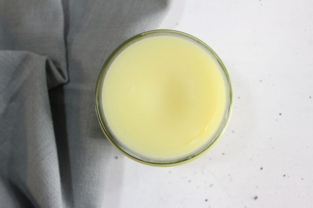 Top view of a glass of pale yellow juice on a white surface. A gray cloth is partially visible on the left side of the image.