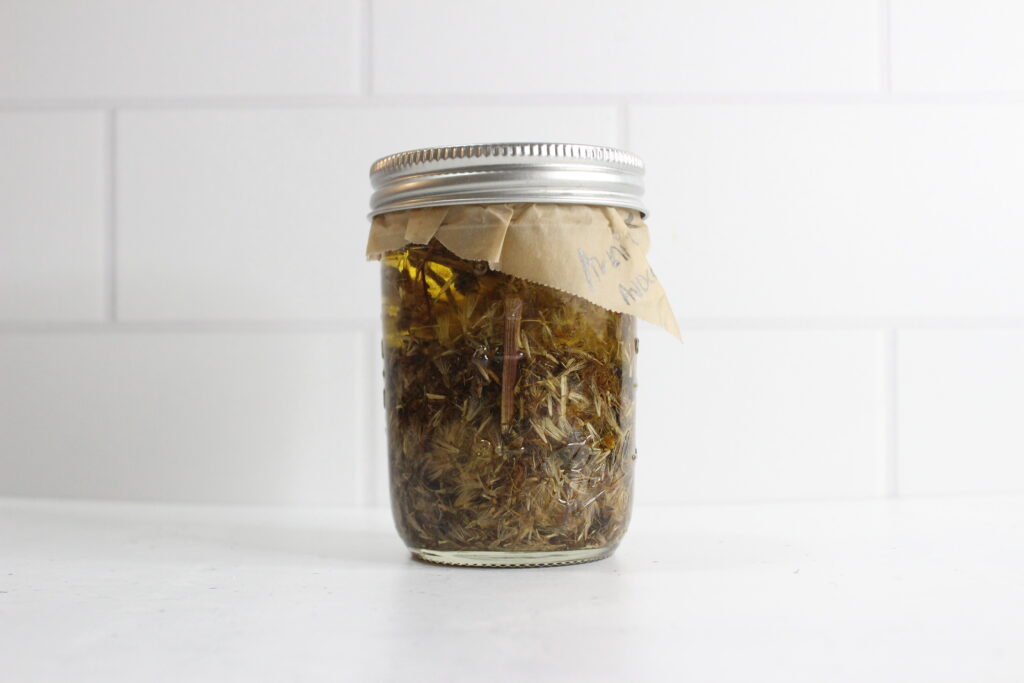A glass jar filled with dried herbs and liquid, sealed with a metal lid. A small piece of brown paper with handwritten text is attached to the jar. The background is a white tiled wall.