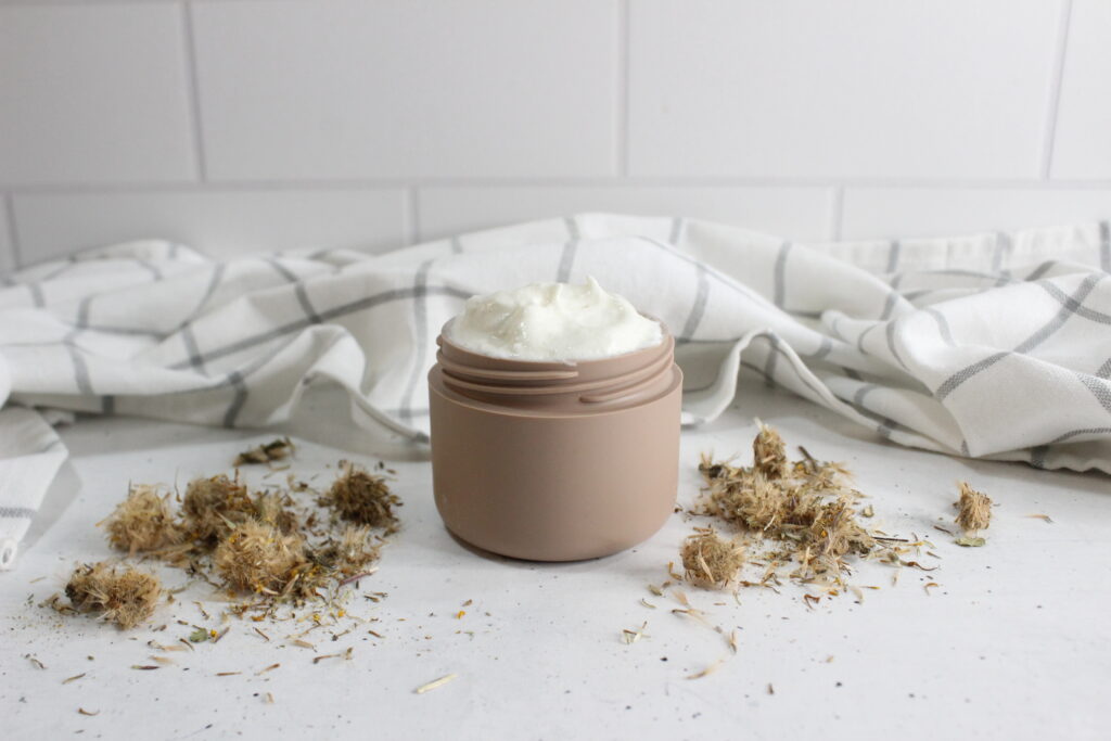 A beige jar of whipped cream sits on a white surface with dried herbs scattered around. In the background, there's a white and gray checkered cloth against a tiled wall.