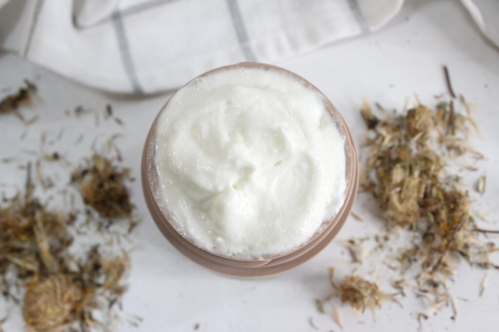 A creamy, frothy cup of hot chocolate or coffee sits on a white surface. Surrounding the cup are small piles of dried herbs or flowers. A white and gray striped cloth is visible in the background.