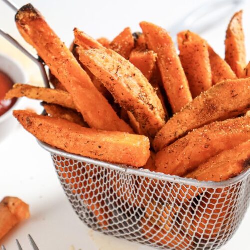 A metal basket filled with seasoned sweet potato fries is on a white surface. A small bowl of red dipping sauce is in the background, partially visible. The fries are golden brown with herbs and spices sprinkled on top.