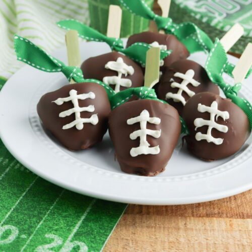 Chocolate-covered strawberries decorated to resemble footballs with white icing on a plate. Green ribbon tops each strawberry, and a football-themed tablecloth is visible in the background.