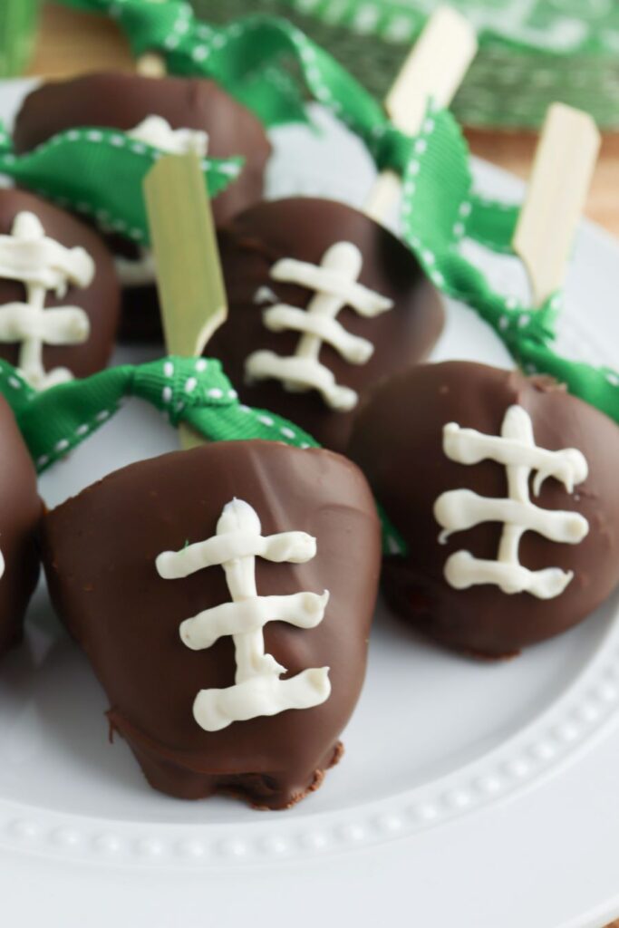 Chocolate-covered strawberries decorated with white icing to resemble footballs, each on a wooden stick with a green ribbon, arranged on a white plate.