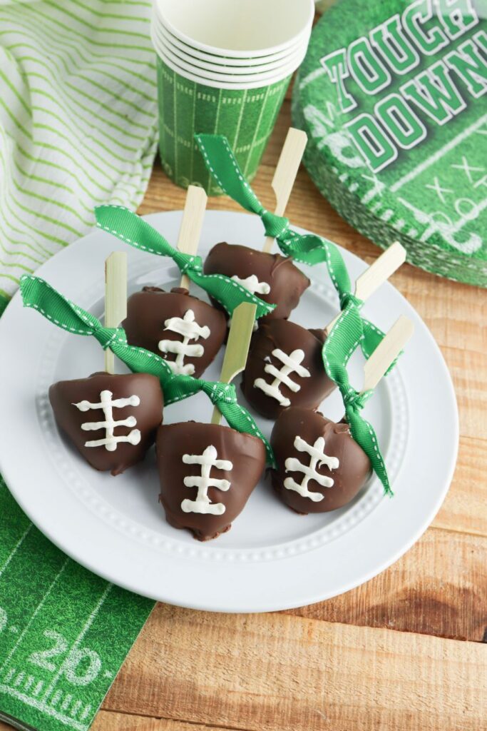 Chocolate-covered treats shaped like footballs on a white plate, each with white icing laces and a wooden stick. Green ribbons are tied to the sticks. Nearby, green-themed napkins and paper cups with a football motif are displayed.
