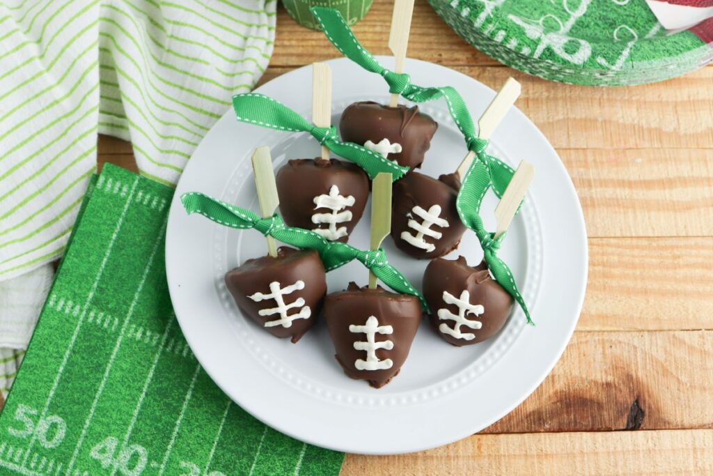 Chocolate-covered treats shaped like footballs, decorated with white icing laces, are arranged on a white plate. Each treat is attached to a wooden stick tied with a green ribbon. The plate is on a wooden table with a green football-themed cloth nearby.