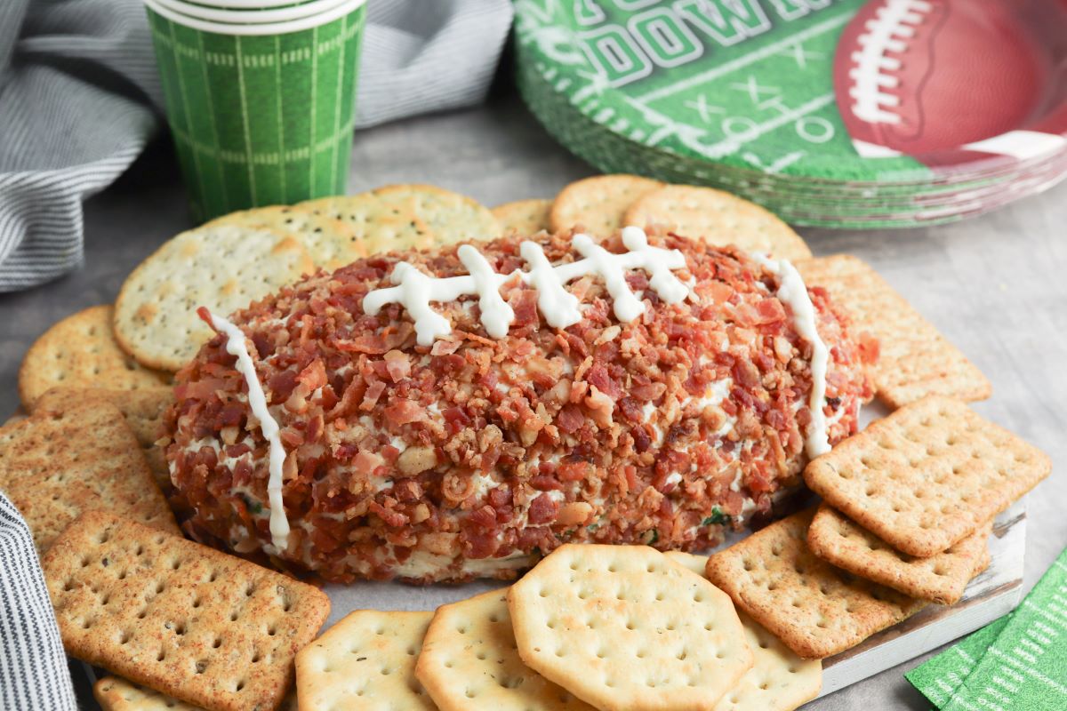 A football-shaped cheese ball covered in bacon bits, decorated with white icing to resemble laces, surrounded by various crackers. Football-themed plates and a green cup are in the background.