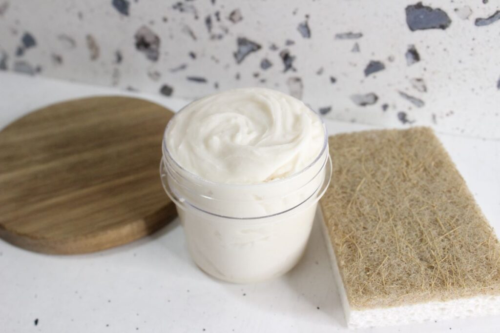 A jar of thick white cream or paste is placed on a countertop with a terrazzo backdrop. A round wooden lid is to the left, and a rectangular sponge with a coarse, brown scrub surface is on the right.
