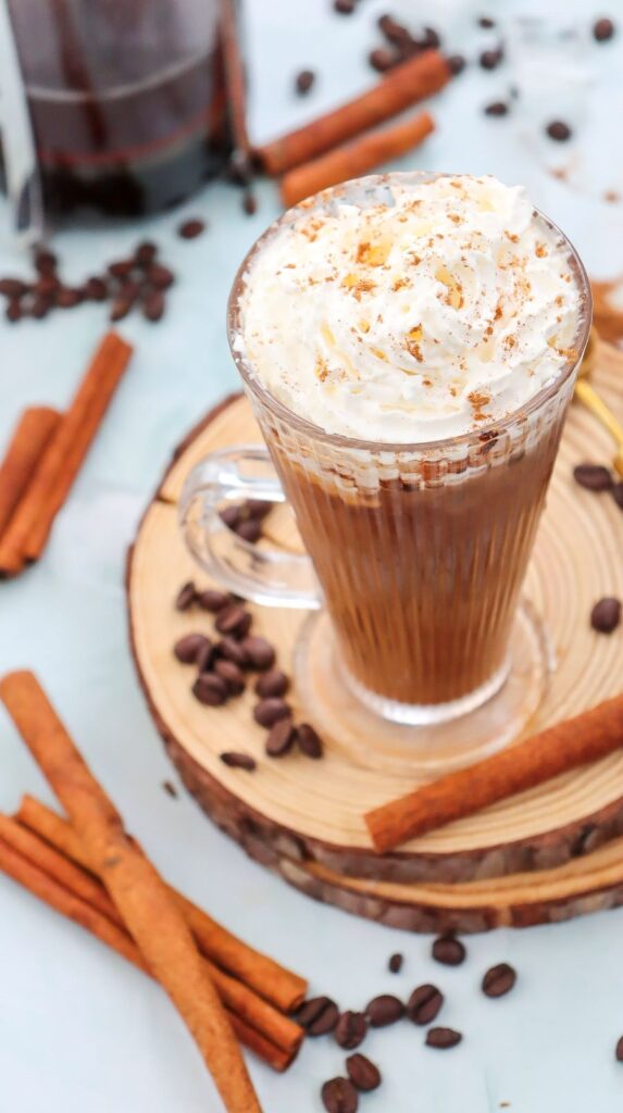 A glass mug of frothy coffee topped with whipped cream and sprinkles sits on a wooden coaster. Cinnamon sticks and coffee beans are scattered around on a light surface.