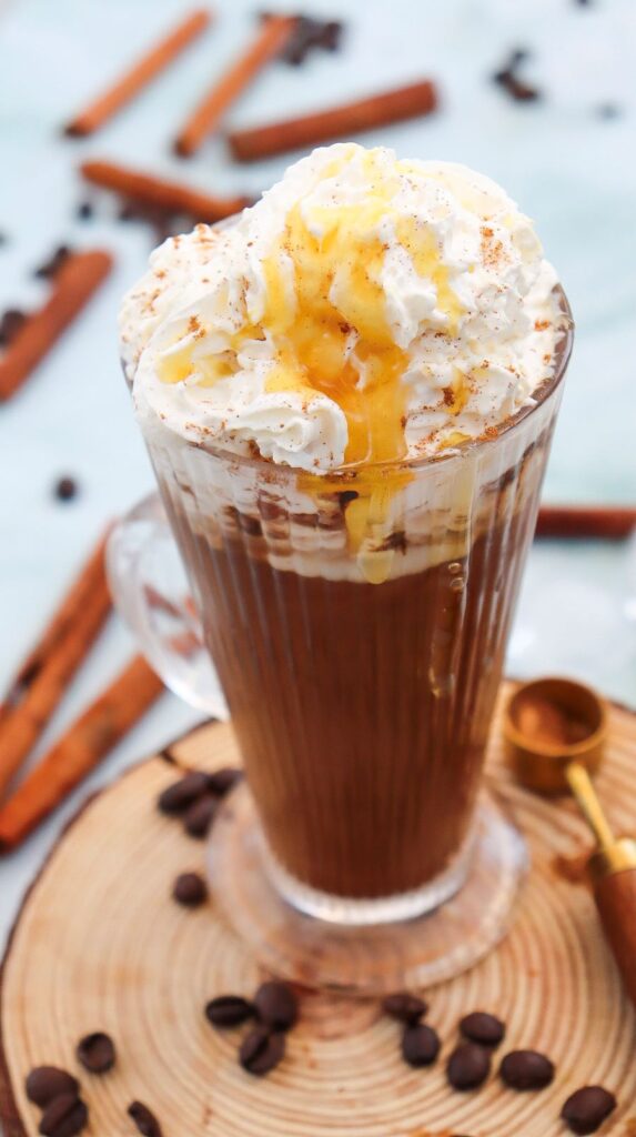 A glass mug of coffee topped with whipped cream and caramel drizzle, placed on a wooden coaster. Cinnamon sticks and coffee beans are scattered around on a light surface.