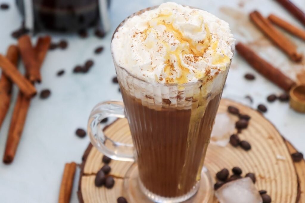 A glass mug of whipped cream-topped coffee sits on a wooden coaster. Drizzled with caramel and sprinkled with cinnamon, the coffee is surrounded by scattered coffee beans and cinnamon sticks on a light surface.